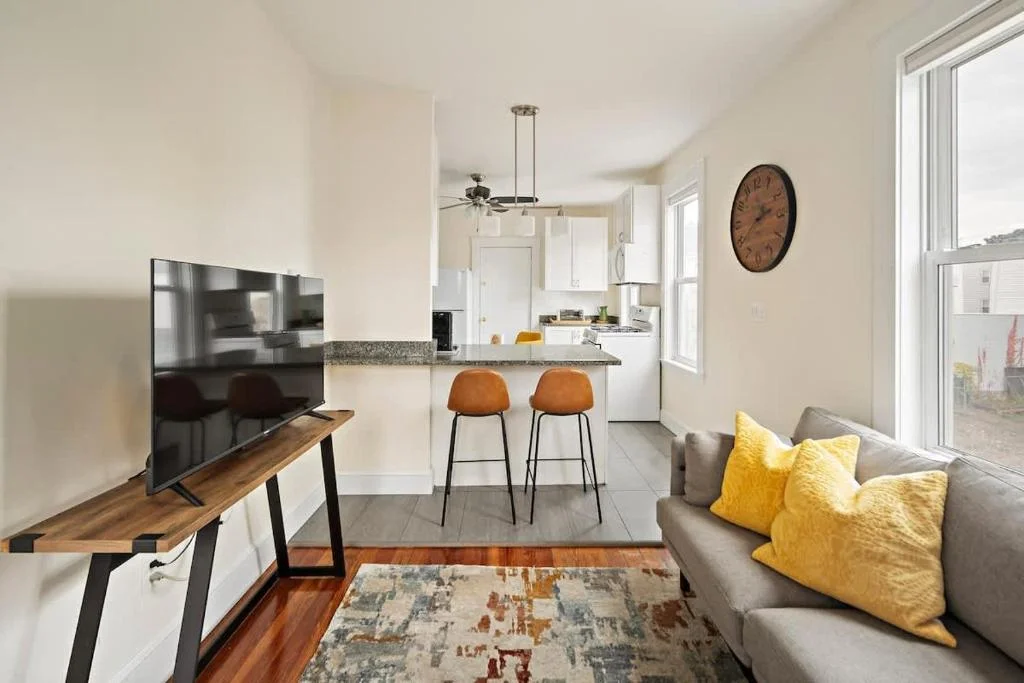 Open-concept living room with grey sofa, yellow pillows, and modern kitchen with orange stools in a Cambridge, MA rental by