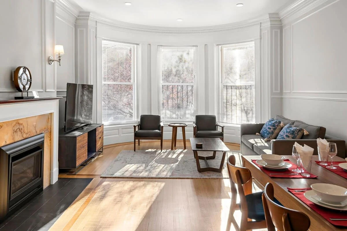 Open-concept living room with bay windows, hardwood floors, and a fireplace beside a dining table in a Boston rental by