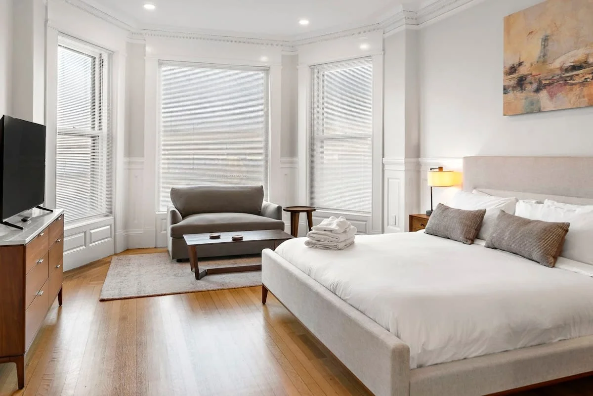 Bedroom with white bedding, folded towels, a gray sofa, and bay windows