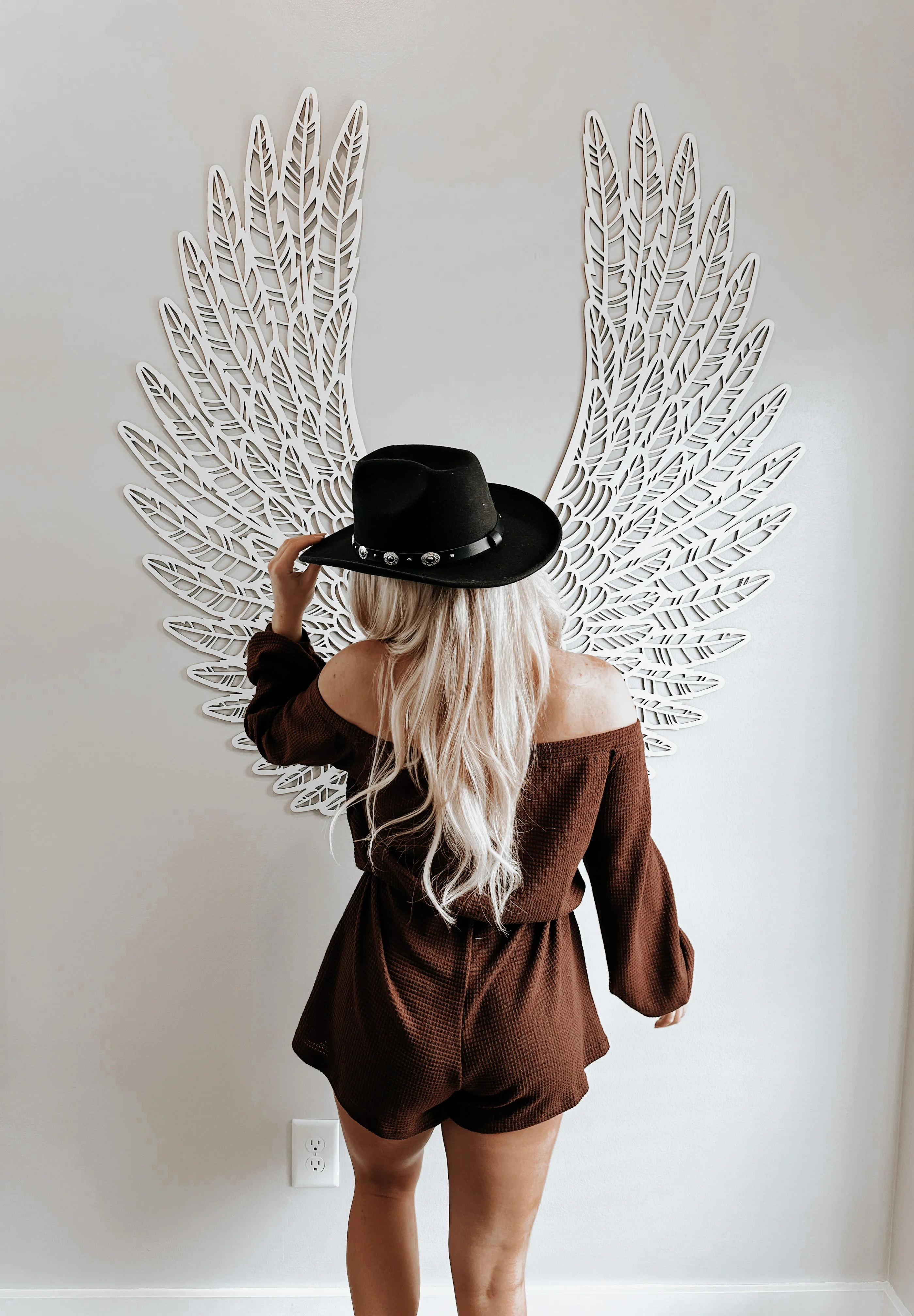 Woman in brown off-shoulder outfit and cowboy hat poses by white feathered wing wall art at Nashville luxury vacation rental