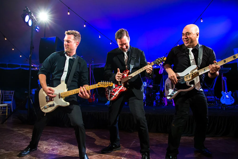 Three musicians perform live wedding processional songs on stage with guitars and bass under blue stage lighting