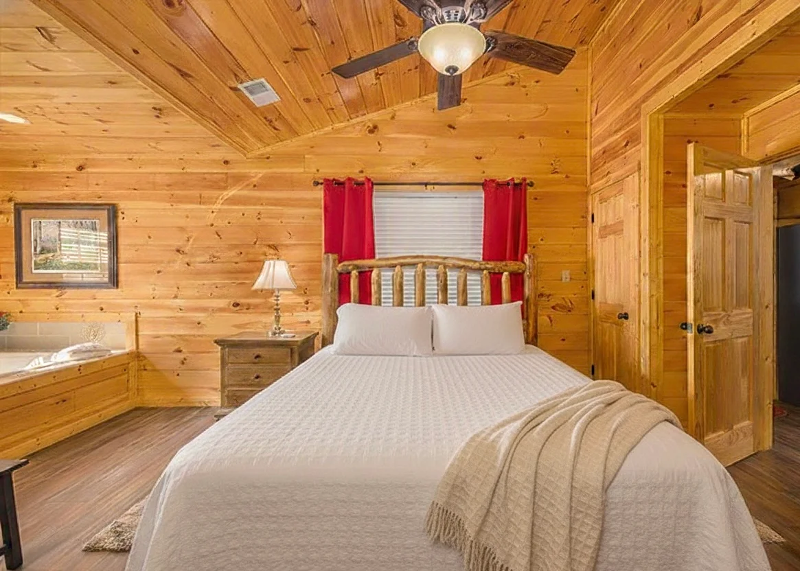 Rustic log cabin bedroom with knotty pine walls, queen bed with red accents, and jacuzzi tub near Sevierville Tennessee