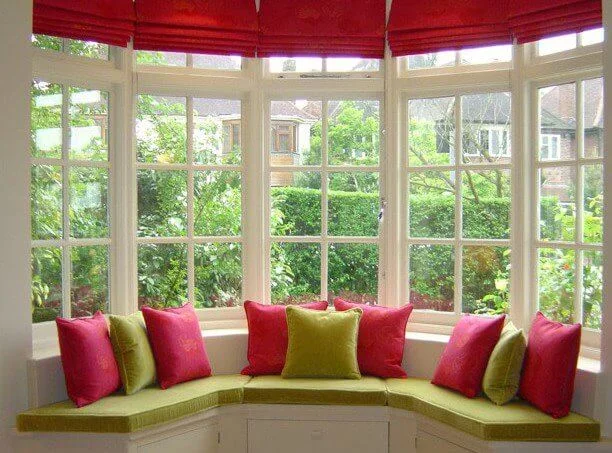 Bright sunroom with curved bay window featuring red roman shades, lime green window seat cushions, and garden views showing