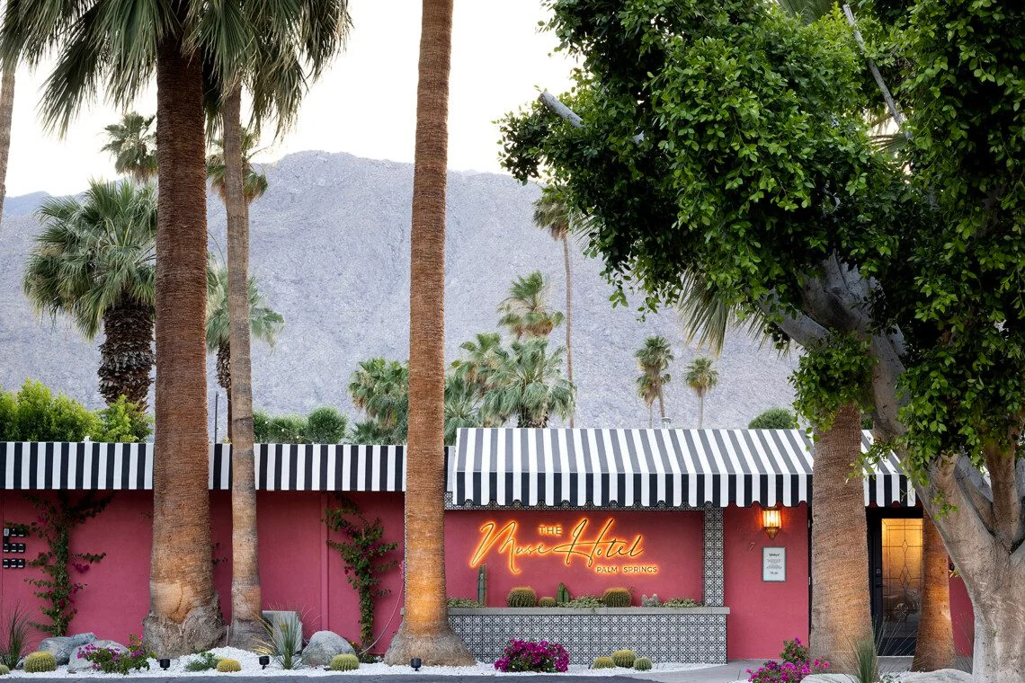 The Muse Hotel's coral-pink mid-century modern facade with palm trees and mountain views, near Palm Springs aerial tramway