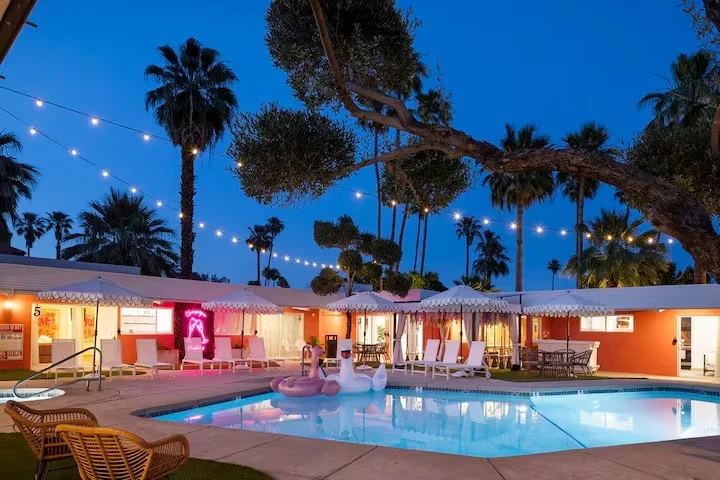 Illuminated hotel pool at dusk with palm trees, string lights, and colorful mid-century modern buildings