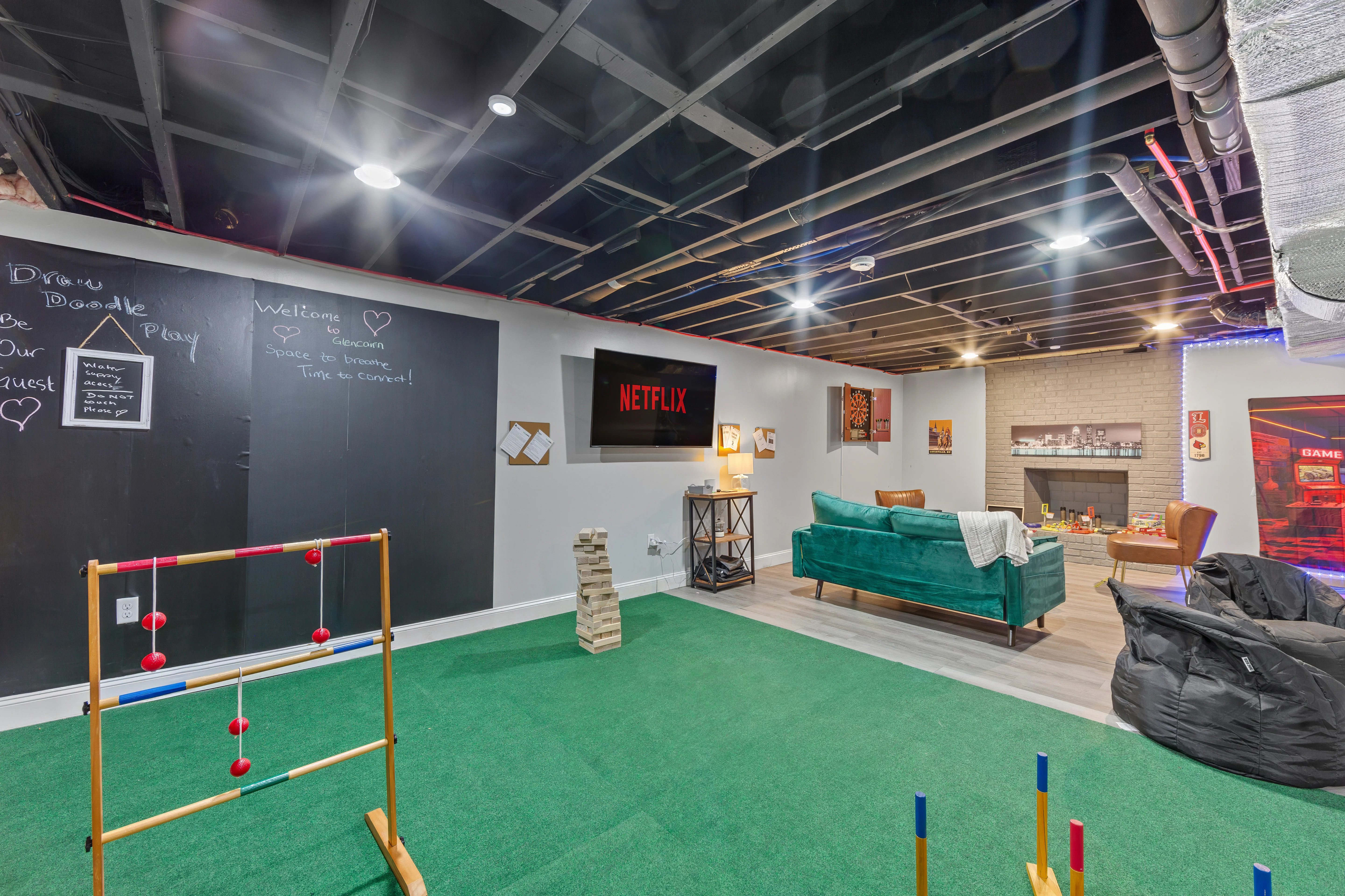 Modern basement game room with green turf, velvet sofa, cornhole, and fireplace at Glencairn Knoll Louisville